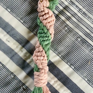 May include: A handmade macrame keychain in a spiral design, featuring alternating sections of pink and green cord. The keychain has a gold-colored clasp and a tassel at the bottom. The background is a striped fabric.