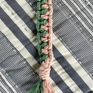 May include: A handmade macrame keychain with a gold-colored clasp. The woven design features alternating sections of green and pink cord, with a tassel at the bottom. The keychain is displayed on a striped fabric.