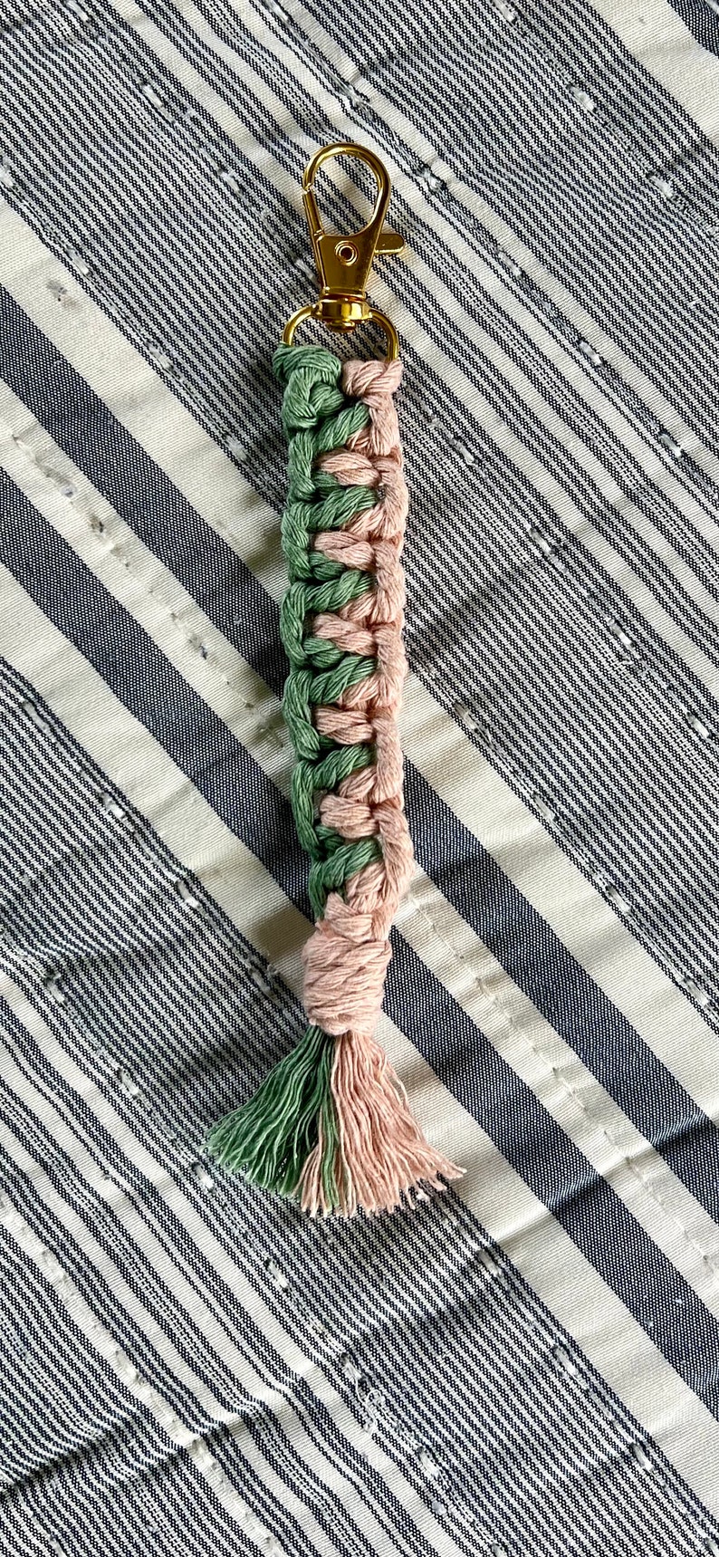May include: A handmade macrame keychain with a gold-colored clasp. The woven design features alternating sections of green and pink cord, with a tassel at the bottom. The keychain is displayed on a striped fabric.