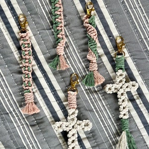 May include: Macrame keychains in various designs, including a cross and braided styles. The keychains feature gold-colored clasps and are made with pink, green, and white cords. The keychains are displayed on a striped background.