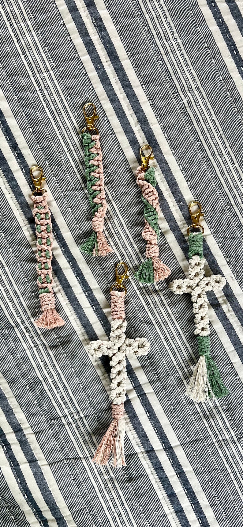 May include: Macrame keychains in various designs, including a cross and braided styles. The keychains feature gold-colored clasps and are made with pink, green, and white cords. The keychains are displayed on a striped background.