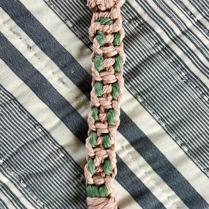 May include: A handmade macrame keychain in a woven pattern, featuring alternating sections of dusty rose and green cord. The keychain has a gold-colored clasp and a tassel at the bottom. The background is a striped fabric.