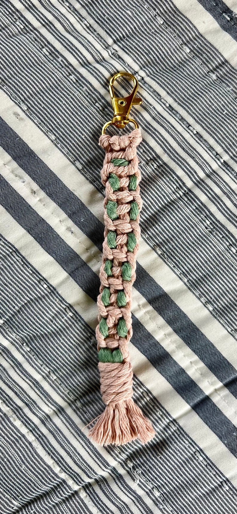 May include: A handmade macrame keychain in a woven pattern, featuring alternating sections of dusty rose and green cord. The keychain has a gold-colored clasp and a tassel at the bottom. The background is a striped fabric.