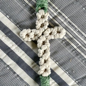 May include: Macrame cross keychain with a gold-colored clasp. The cross is made of off-white macrame knots, with green macrame accents and a tassel. The background is a striped fabric.