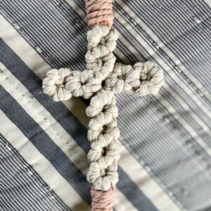 May include: Macrame cross keychain with a gold-colored clasp. The cross is made of off-white and pink knotted cord, with a tassel at the bottom. The cross is displayed on a striped fabric background.