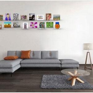 May include: A gray sectional sofa with two orange throw pillows sits in front of a white wall with a gallery wall of framed artwork. The artwork includes a variety of styles and colors, including abstract, landscape, and portrait.