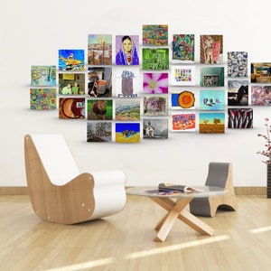 May include: A white wall with a gallery wall of framed artwork. The artwork is a mix of styles and colors, including landscapes, portraits, and abstract art. There is a white chair and a wooden table in the foreground.