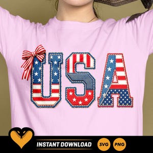 Könnte beinhalten: Ein hellrosa T-Shirt mit dem Schriftzug USA in einem patriotischen Flaggenmuster in Rot, Weiß und Blau mit Sternen und Streifen. Eine rot-weiß gestreifte Schleife ist an der linken Seite des Buchstabens U auf der Vorderseite befestigt.