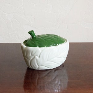 May include: A white ceramic sugar bowl with a green leaf-shaped lid. The bowl is decorated with a leaf pattern.