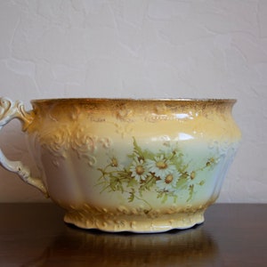 May include: A large, white ceramic bowl with a gold rim and floral design. The bowl has a single handle and is decorated with a pattern of white daisies and green leaves.