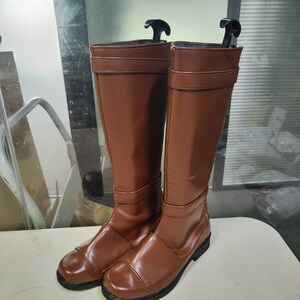 May include: A pair of brown leather knee-high boots with a buckle detail on the front. The boots have a black sole and a pointed toe.