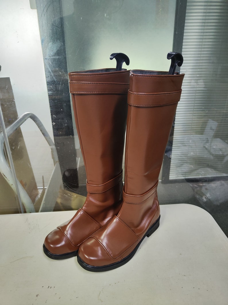 May include: A pair of brown leather knee-high boots with a buckle detail on the front. The boots have a black sole and a pointed toe.