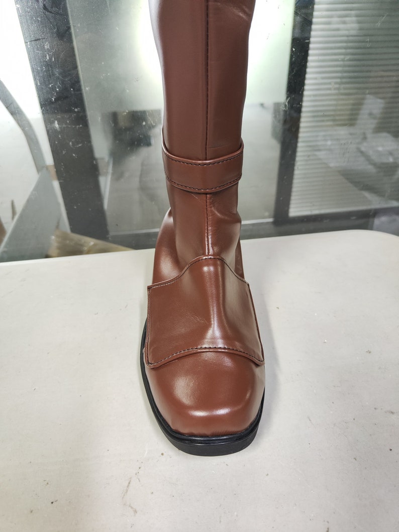 May include: Brown leather knee-high boots with a buckle strap and a square toe.