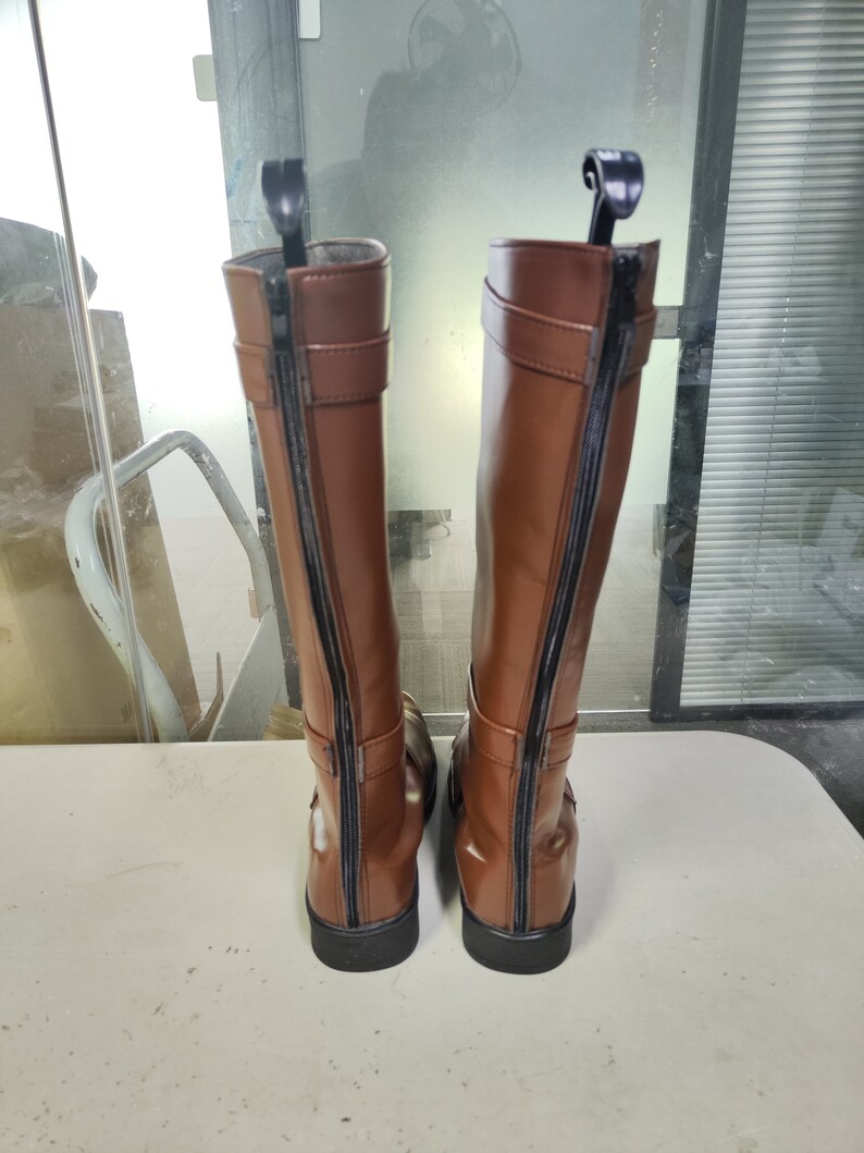 May include: A pair of brown leather knee-high boots with black zips on the back. The boots have a buckle detail on the side.