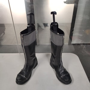 May include: A pair of black leather boots with a zipper up the front and silver studs on the toe. The boots have a gray fabric lining and are on a white surface.