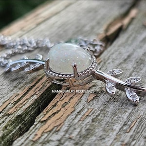 Peut inclure: Un collier argenté avec un grand pendentif opalescent ovale. Le pendentif est serti dans un cadre argenté décoratif, avec des accents en forme de feuilles sur la chaîne. Le texte "MAMA'S MILKY KEEPSAKES" est visible.