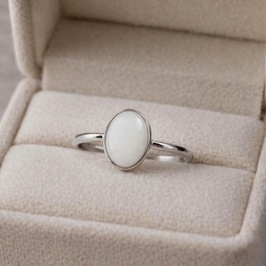 May include: A silver ring with an oval, white gemstone set in a bezel. The ring is displayed in a beige velvet ring box, with the ring centered in the frame. The ring's band is thin and polished.