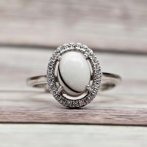 May include: A silver ring featuring a white oval gemstone set within a halo of smaller clear gemstones.