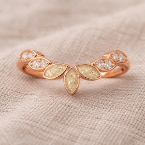 May include: A rose gold ring with a leaf-like design. The ring features clear, round gemstones and marquise-shaped sections filled with a creamy, speckled material. The ring is set against a neutral, textured background.