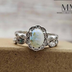 Pu&ograve; includere: Un anello d'argento con una grande pietra ovale di colore bianco latte circondata da piccole pietre trasparenti. L'anello &egrave; su una superficie marrone. MM Mama's Milky Keepsakes