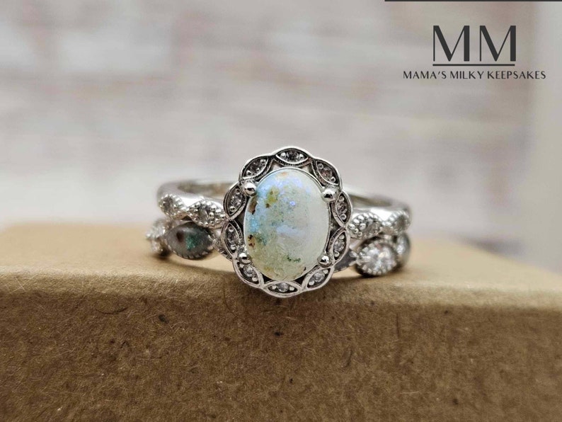 Pu&ograve; includere: Un anello d'argento con una grande pietra ovale di colore bianco latte circondata da piccole pietre trasparenti. L'anello &egrave; su una superficie marrone. MM Mama's Milky Keepsakes