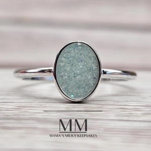 Puede incluir: Un anillo de plata con una piedra ovalada de color azul claro llena de purpurina. El anillo está sobre una superficie de madera clara. El texto "MM Mama's Milky Keepsakes" está debajo del anillo.