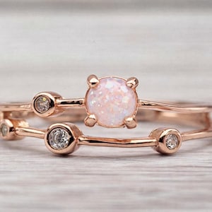 May include: A rose gold ring set featuring a double band design. The top ring has a round, light pink opal center stone. The lower band and the top band have small, clear accent stones.