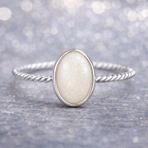 May include: A silver ring with a cream-colored oval stone. The band has a twisted rope design. The ring is set against a blurred, sparkly background, creating a soft, elegant aesthetic.