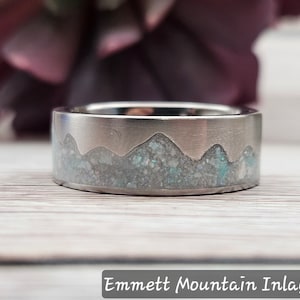 May include: A silver ring with a mountain range inlay made of white and blue speckled material. The ring is on a light wood surface. Emmett Mountain Inlay.