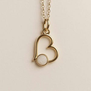May include: A gold-colored pendant necklace featuring a heart-shaped design with a circular, white stone at the bottom. The pendant hangs from a delicate gold chain, creating a simple and elegant piece of jewelry.