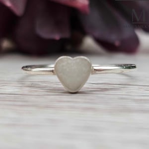 May include: A silver ring with a white heart-shaped stone. The ring is on a white wooden surface.