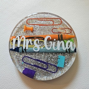 May include: A round, clear resin coaster with a silver glitter background. The coaster features a variety of school supplies, including paper clips, push pins, crayons, and a pencil. The text "Mrs. Gina" is written in white letters on the coaster.