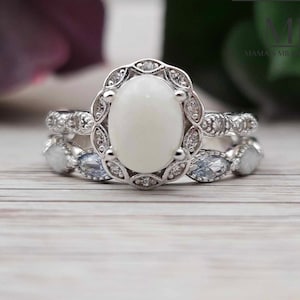 Pu&ograve; includere: Un anello d'argento con un grande opale bianco ovale circondato da pietre pi&ugrave; piccole e trasparenti. L'anello ha un design delicato di ispirazione vintage.