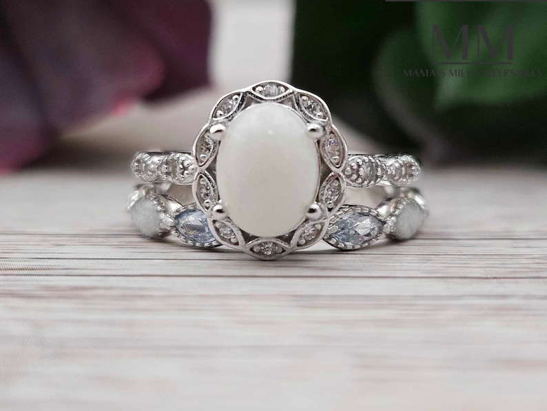 Pu&ograve; includere: Un anello d'argento con un grande opale bianco ovale circondato da pietre pi&ugrave; piccole e trasparenti. L'anello ha un design delicato di ispirazione vintage.