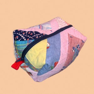 May include: A colourful patchwork pouch with a navy blue zip and a red pull tab. The fabrics used include floral patterns, polka dots, and solid colours. The pouch is made from various fabrics.