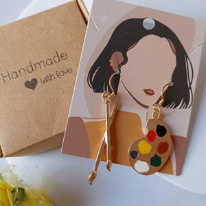 May include: Gold-coloured earrings with a paintbrush and artist's palette design. The palette has various paint colours. The earrings are displayed on a card with a woman's illustration. A small brown box with "Handmade with love" is also in the image.