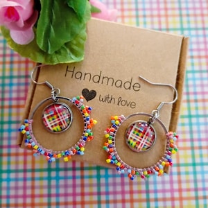 Puede incluir: Un par de pendientes de aro hechos a mano con un diseño de cuadros coloridos. Los pendientes tienen un marco de metal plateado y están adornados con pequeñas cuentas multicolores. Se exhiben en una caja de cartón marrón con el texto "Handmade with love."
