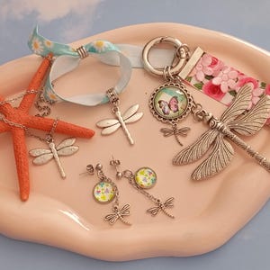 Op de afbeelding: Een collectie sieraden met zilverkleurige libellebedels, waaronder een ketting met een oranje zeester, een lintarmband, een sleutelhanger met bloemenpatroon en een paar oorbellen met kleine ronde bloemencabochons.