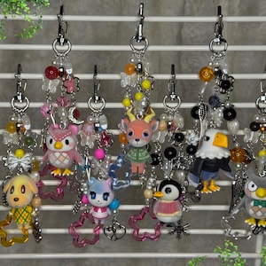 Peut inclure: Une collection de porte-clés de personnages avec des accents de perles et des fermoirs mousquetons. Les porte-clés présentent divers personnages d'animaux, dont un hibou, un chien, un cerf, un pingouin et un aigle. Chaque porte-clés a un design et une palette de couleurs uniques.