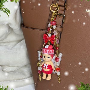 May include: A decorative keychain featuring a small doll dressed in red with a matching hat and wings. The keychain includes red beads, a red bow, and a gold-colored clasp, attached to a brown handbag.