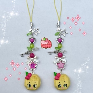 May include: Two phone charms featuring a yellow waffle character with a raspberry on top, a pink star bead, a white bow, a silver key charm, green and pink beads, and a silver star charm, all attached to a yellow cord with a small metal clasp.