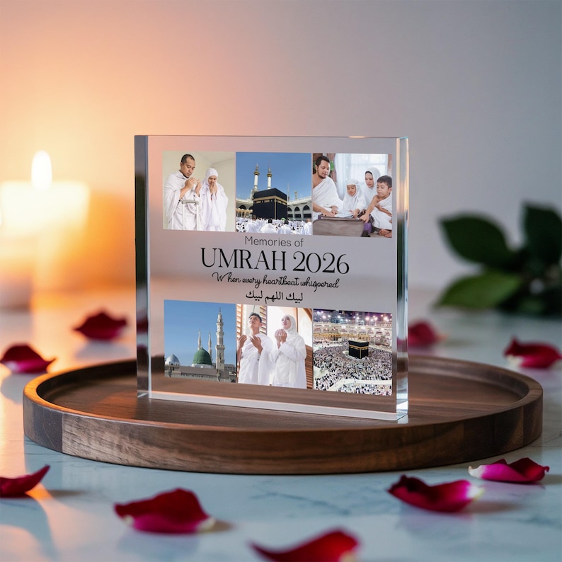 May include: A clear acrylic photo block with the text "Memories of Umrah 2026" and images of religious sites and people. The block sits on a wooden tray, surrounded by red rose petals and a lit candle.