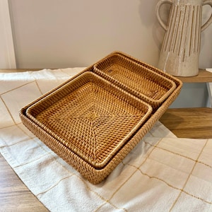 May include: Set of three rectangular woven rattan trays in varying sizes. The trays are a light brown color and have a textured surface. The trays are nested together on a white and gold patterned tablecloth.
