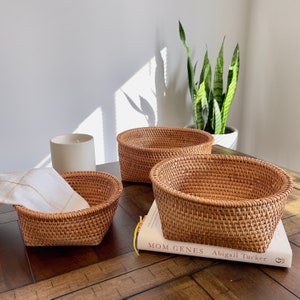 May include: Three woven rattan baskets of varying sizes sit on a dark wood surface. The baskets are a warm brown color and have a textured, open weave design. A white candle and a book titled "MOM GENES" are also visible.