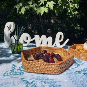 May include: A hexagonal woven rattan tray filled with dark red cherries sits on a blue and white patterned tablecloth. A second tray with pastries is visible. A green glass diffuser bottle and a white "home" sign are in the background.