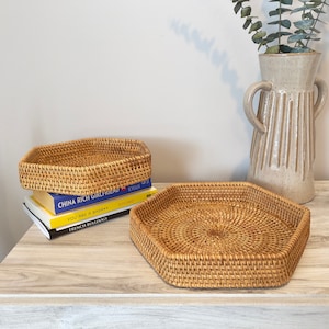 May include: Two woven rattan trays in a natural tan color. One tray is hexagonal, and the other is a smaller, rectangular shape. The trays are on a light-colored wooden surface, with books and a decorative vase in the background.