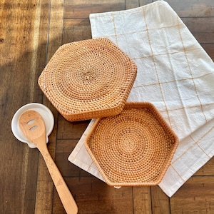 May include: A wooden honey dipper and spoon rest set with two woven rattan trivets and a white and gold checkered kitchen towel. The spoon has a honeycomb design and bee graphic. The trivets are hexagonal and square.