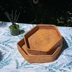 Könnte beinhalten: Zwei hellbraune, gewebte Rattan-Tabletts in sechseckiger Form, die ineinander verschachtelt sind. Sie liegen auf einer weißen Tischdecke mit blauem Blättermuster. Im Hintergrund ist eine grüne Glasdiffusorflasche mit Schilf zu sehen.