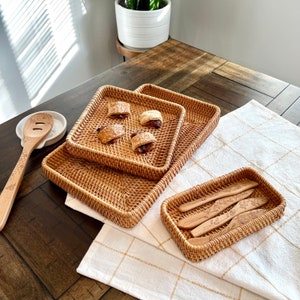 Puede incluir: Bandejas de servicio de ratán tejido y una pequeña cesta rectangular con cuchillos de mantequilla de madera. Los utensilios de madera tienen un diseño floral. Las bandejas están vacías y la cesta contiene cuatro cuchillos. Una cuchara de madera descansa sobre un reposacucharas de cerámica blanca.