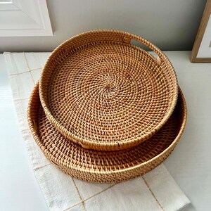 Op de afbeelding: Twee ronde, geweven rotan trays in natuurlijke tinten. De trays zijn genest, waarbij de ene iets groter is dan de andere. De trays hebben een spiraalvormig weefpatroon en zijn geplaatst op een wit oppervlak met een cr&egrave;mekleurige, geblokte doek.